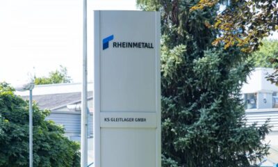 Tall white outdoor sign for RHEINMETALL with a blue logo, displaying KS GLEITLAGER GMBH, placed near trees and a building exterior