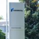 Tall white outdoor sign for RHEINMETALL with a blue logo, displaying KS GLEITLAGER GMBH, placed near trees and a building exterior