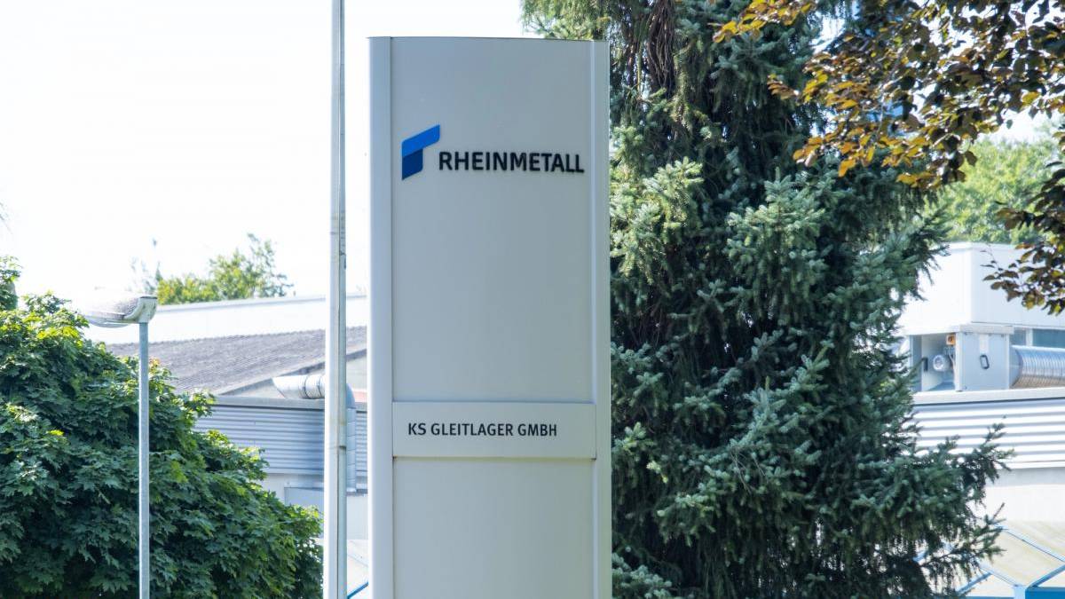 Tall white outdoor sign for RHEINMETALL with a blue logo, displaying KS GLEITLAGER GMBH, placed near trees and a building exterior