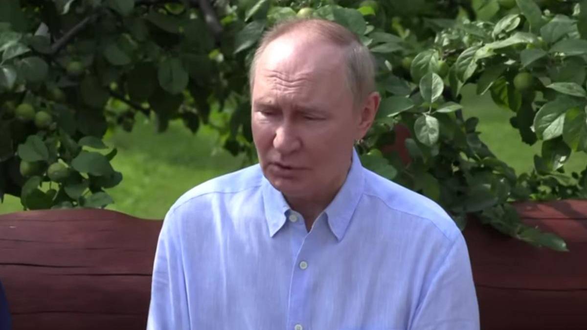 A man in a light blue shirt sits on a wooden bench outdoors, with green foliage in the background.
