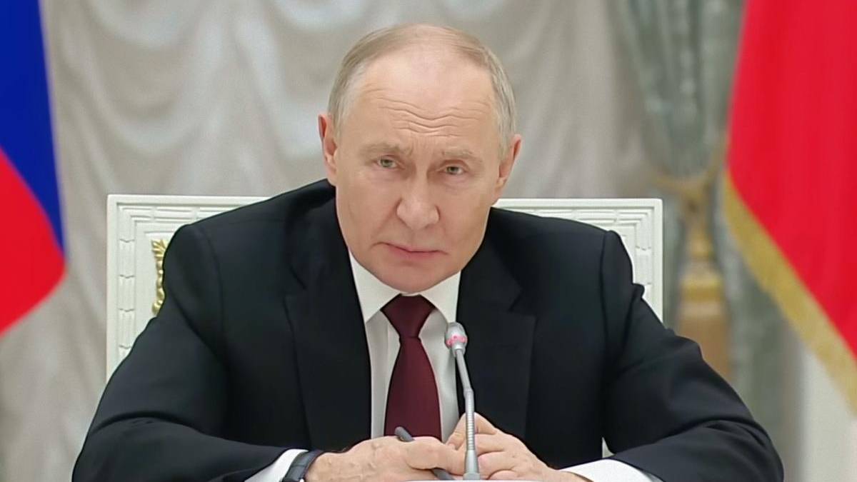 Man in a dark suit and red tie sits at a conference table, speaking into a microphone at a formal event.