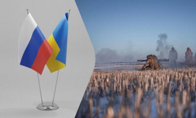 Two national flags, Russia and Ukraine, mounted on a stand, with a distant battlefield scene on the right showing soldiers in a smoky field.