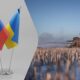 Two national flags, Russia and Ukraine, mounted on a stand, with a distant battlefield scene on the right showing soldiers in a smoky field.