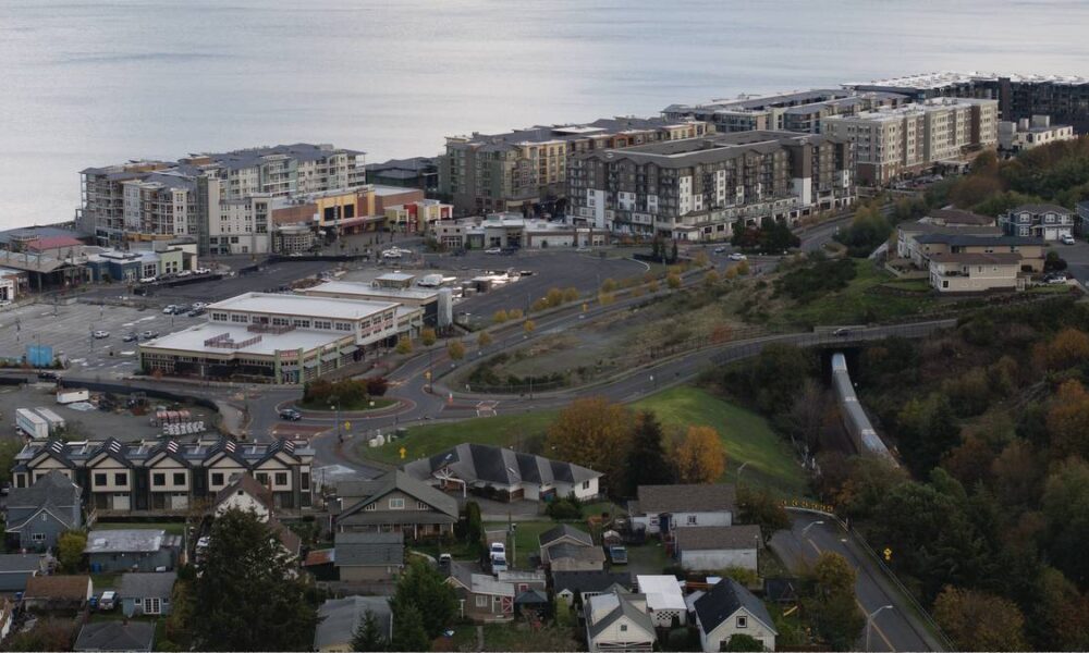 Aerial view of a coastal town with apartment buildings along the shoreline, a shopping plaza, and winding roads.