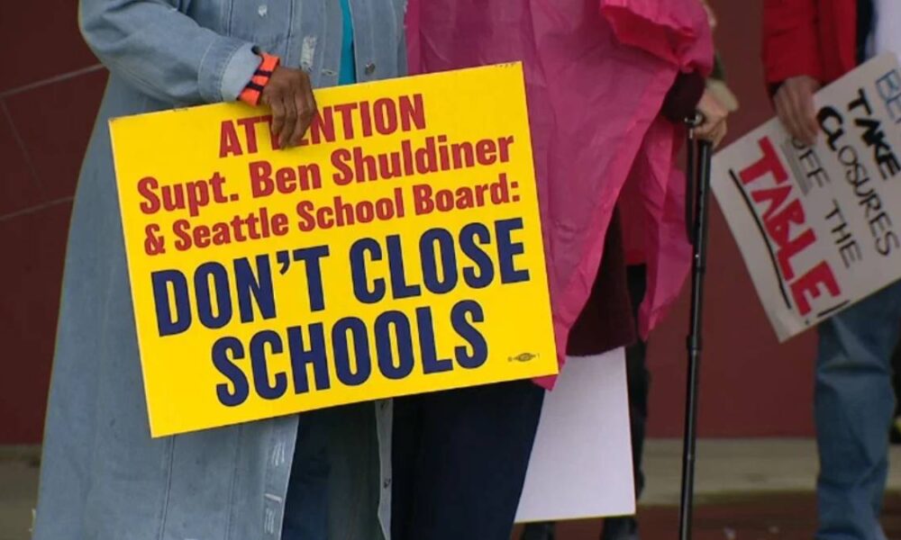 Bright yellow protest sign reading 'DONT CLOSE SCHOOLS' held by a person in a denim jacket, with others and signs in the background.