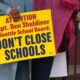 Bright yellow protest sign reading 'DONT CLOSE SCHOOLS' held by a person in a denim jacket, with others and signs in the background.