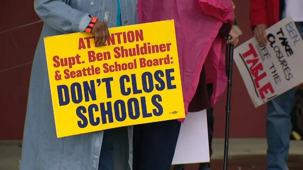 Bright yellow protest sign reading 'DONT CLOSE SCHOOLS' held by a person in a denim jacket, with others and signs in the background.
