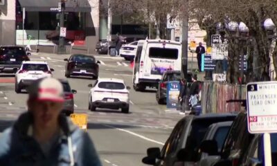 Busy city street with cars and a white bus, blurred cyclist in foreground on a sunny day.