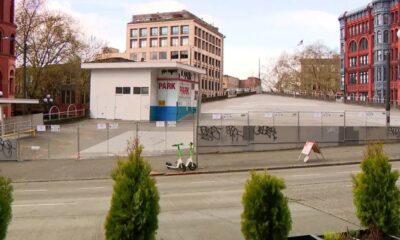 City street with a fenced, graffiti-covered vacant lot, a white kiosk labeled 'PARK', and red brick buildings in the background.
