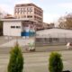 City street with a fenced, graffiti-covered vacant lot, a white kiosk labeled 'PARK', and red brick buildings in the background.