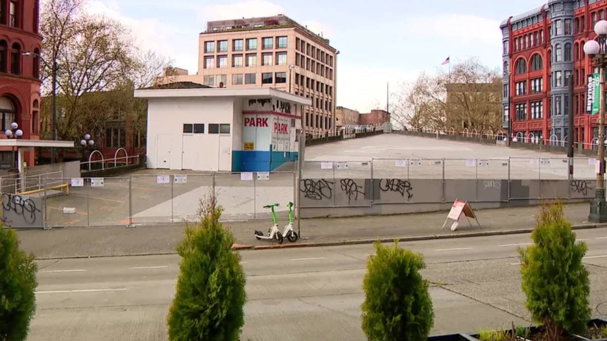City street with a fenced, graffiti-covered vacant lot, a white kiosk labeled 'PARK', and red brick buildings in the background.