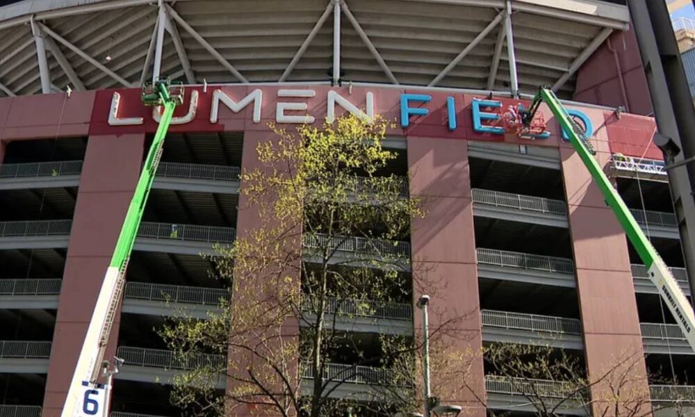 Two green boom lifts install the LUMEN FIELD sign on a stadium facade, with workers on the lifts and balconies visible behind.
