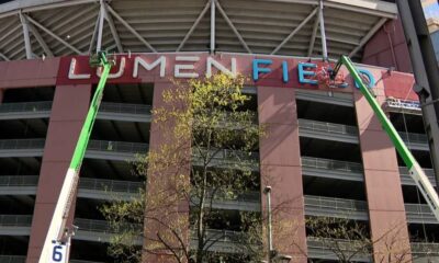 Two green boom lifts install the LUMEN FIELD sign on a stadium facade, with workers on the lifts and balconies visible behind.