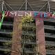 Two green boom lifts install the LUMEN FIELD sign on a stadium facade, with workers on the lifts and balconies visible behind.