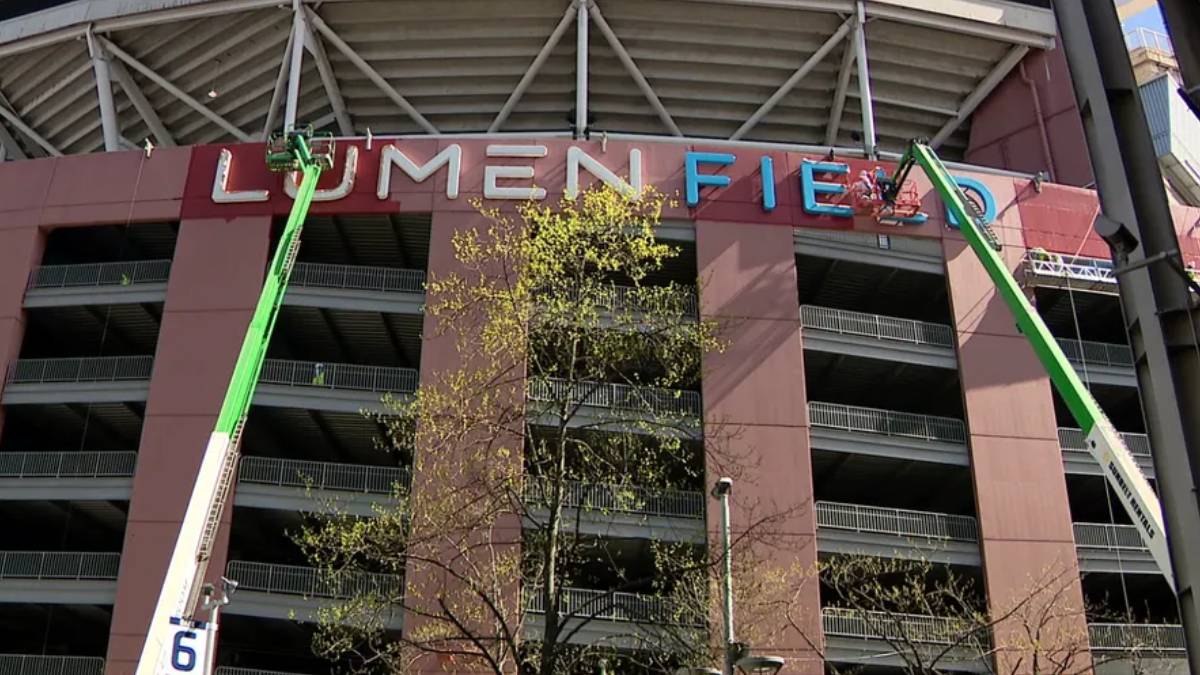 Two green boom lifts install the LUMEN FIELD sign on a stadium facade, with workers on the lifts and balconies visible behind.
