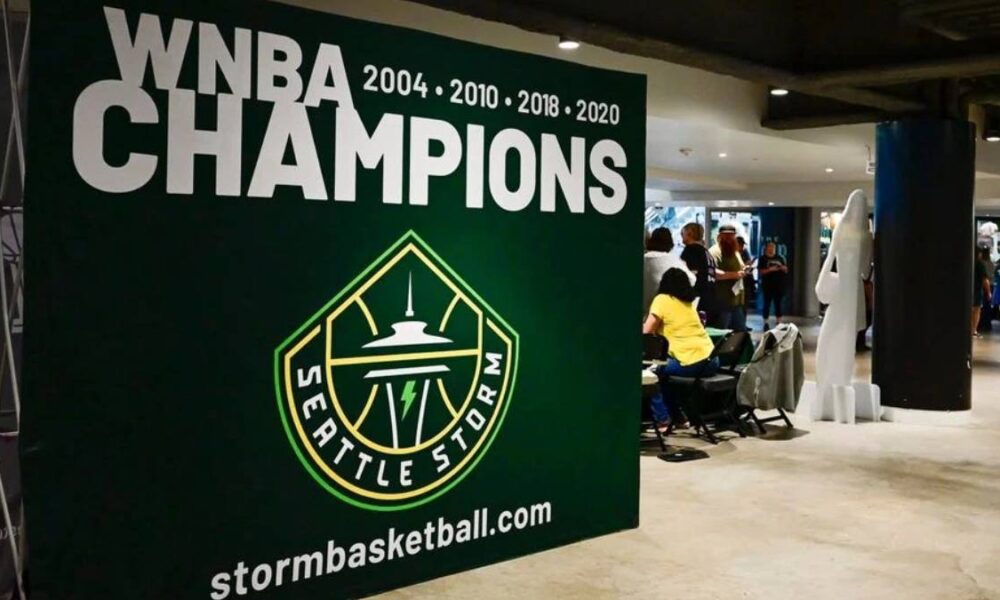 Large green banner reading 'WNBA CHAMPIONS' with years and the Seattle Storm logo, plus stormbasketball.com, in an indoor event space with people nearby.