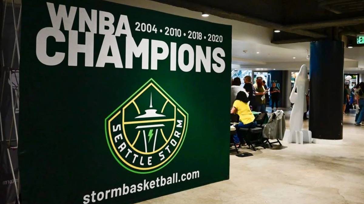 Large green banner reading 'WNBA CHAMPIONS' with years and the Seattle Storm logo, plus stormbasketball.com, in an indoor event space with people nearby.