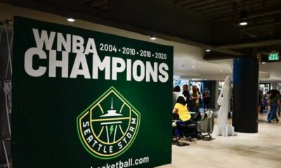 Large green banner reading 'WNBA CHAMPIONS' with Seattle Storm logo, celebrating championships (2004–2010–2018–2020). Inside venue with people nearby.