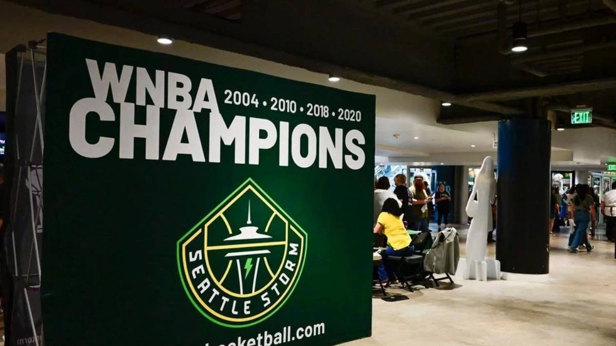 Large green banner reading 'WNBA CHAMPIONS' with Seattle Storm logo, celebrating championships (2004–2010–2018–2020). Inside venue with people nearby.