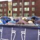 Row of colorful tents and tarps pitched behind a chain-link fence, with portable toilets on the left and a brown multi-story apartment building in the background.