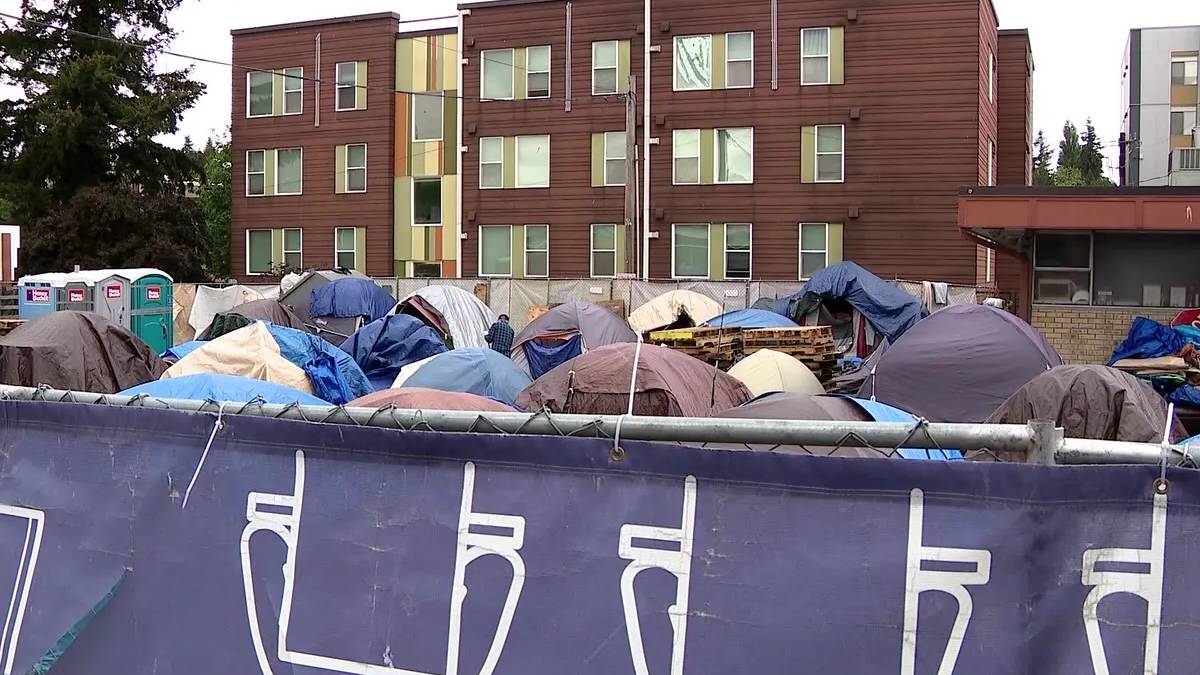 Row of colorful tents and tarps pitched behind a chain-link fence, with portable toilets on the left and a brown multi-story apartment building in the background.