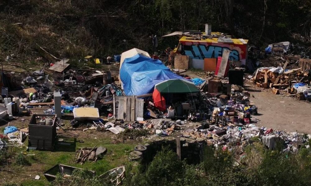 A cluttered outdoor encampment with tents, blue tarps, and piles of discarded furniture and debris across a dusty lot.
