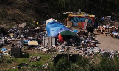 A cluttered outdoor encampment with tents, blue tarps, and piles of discarded furniture and debris across a dusty lot.