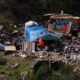 A cluttered outdoor encampment with tents, blue tarps, and piles of discarded furniture and debris across a dusty lot.