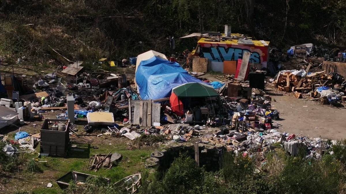 A cluttered outdoor encampment with tents, blue tarps, and piles of discarded furniture and debris across a dusty lot.