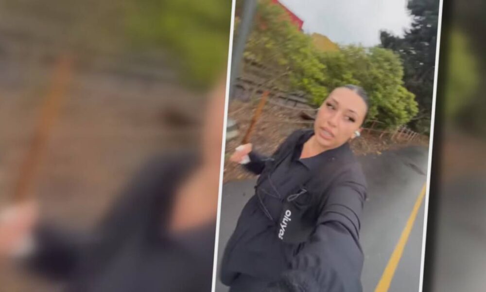 Woman in a black jacket taking a selfie on a residential street, with trees and a stone wall behind her.