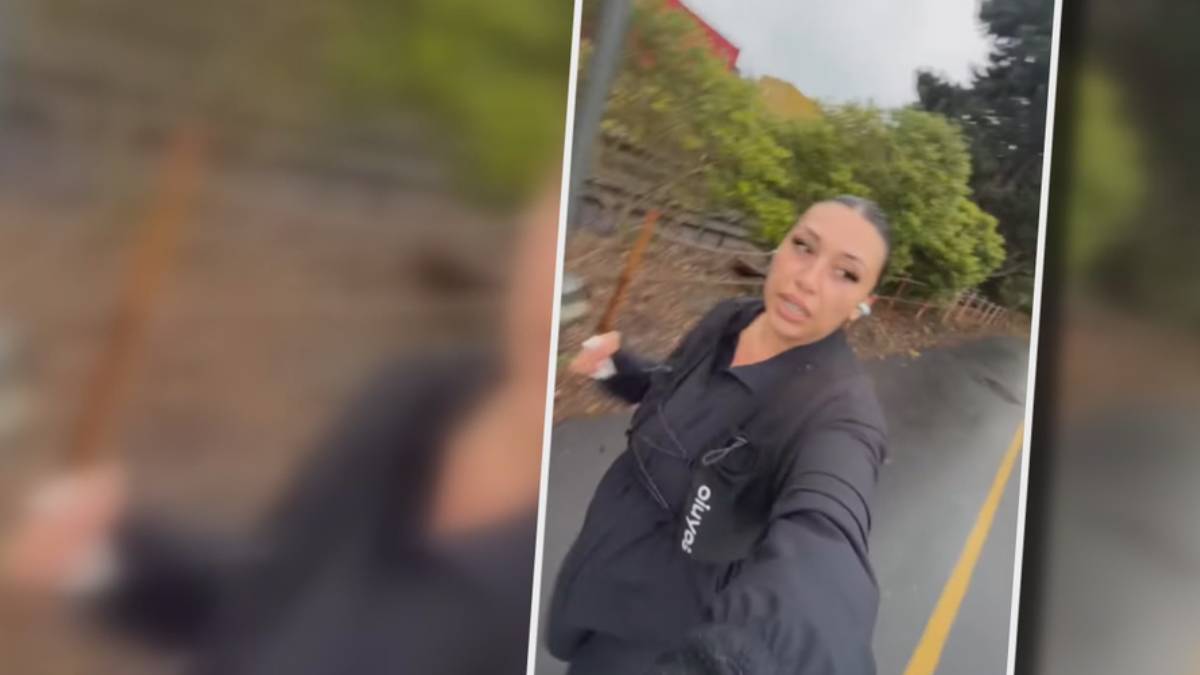 Woman in a black jacket taking a selfie on a residential street, with trees and a stone wall behind her.