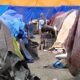 Row of tents and blue tarps forming a makeshift campsite, with scattered belongings on the ground and a person standing in the distance near a cart or stroller.