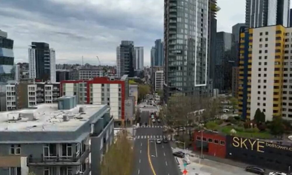 Urban city street scene with tall glass towers, mid-rise buildings, and a wide road leading into the distance.