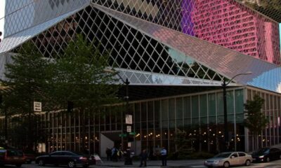 Colorful glass-clad modern building with diagonal panels on a city street, surrounded by cars and pedestrians.