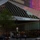 Colorful glass-clad modern building with diagonal panels on a city street, surrounded by cars and pedestrians.