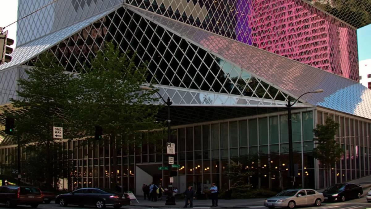 Colorful glass-clad modern building with diagonal panels on a city street, surrounded by cars and pedestrians.