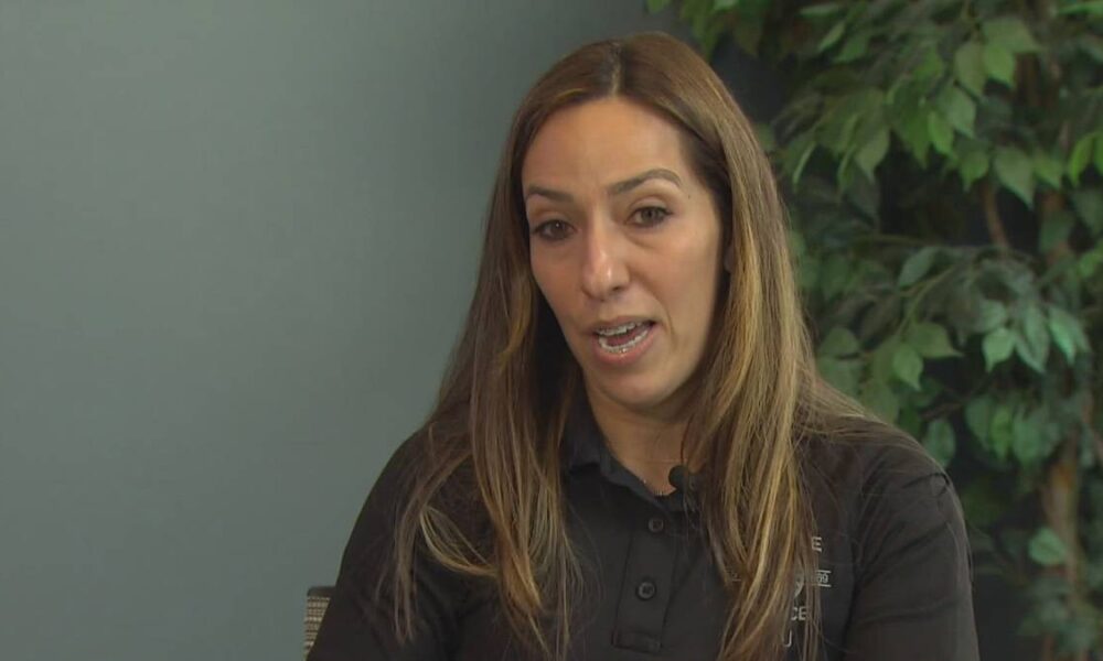 Close-up of a woman with long brown hair wearing a black polo shirt speaking, with green plants in the background (interview setting).