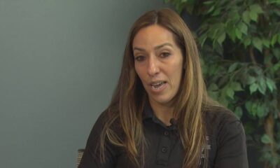 Close-up of a woman with long brown hair wearing a black polo shirt speaking, with green plants in the background (interview setting).