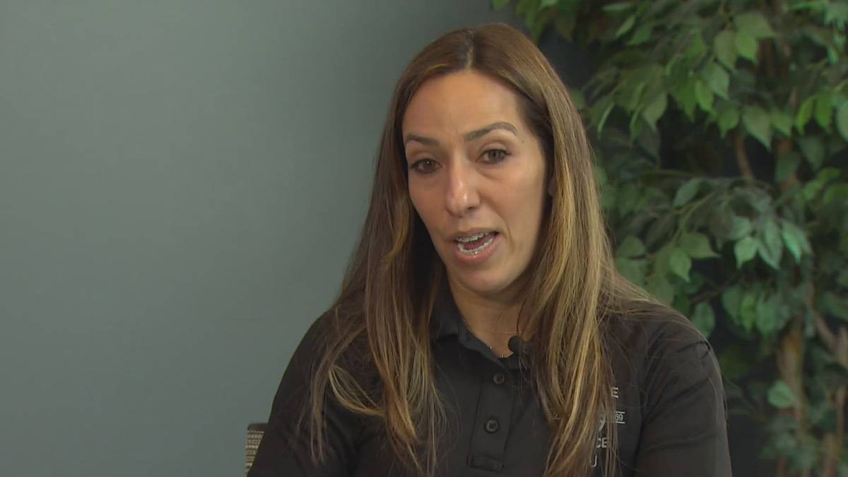 Close-up of a woman with long brown hair wearing a black polo shirt speaking, with green plants in the background (interview setting).