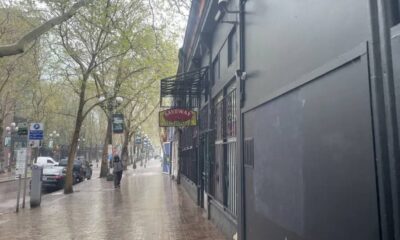 A rainy city sidewalk with wet bricks, a person walking away under trees, and a dark building with a sign hanging from a balcony on the right.