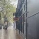 A rainy city sidewalk with wet bricks, a person walking away under trees, and a dark building with a sign hanging from a balcony on the right.