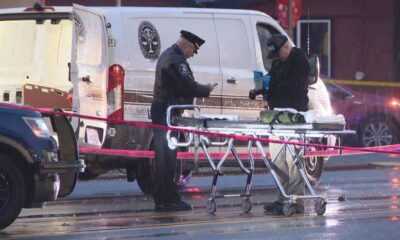 Emergency responders stand beside a wheeled stretcher at a taped-off crime scene, with a white van in the background and a police vehicle nearby.
