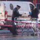 Emergency responders stand beside a wheeled stretcher at a taped-off crime scene, with a white van in the background and a police vehicle nearby.