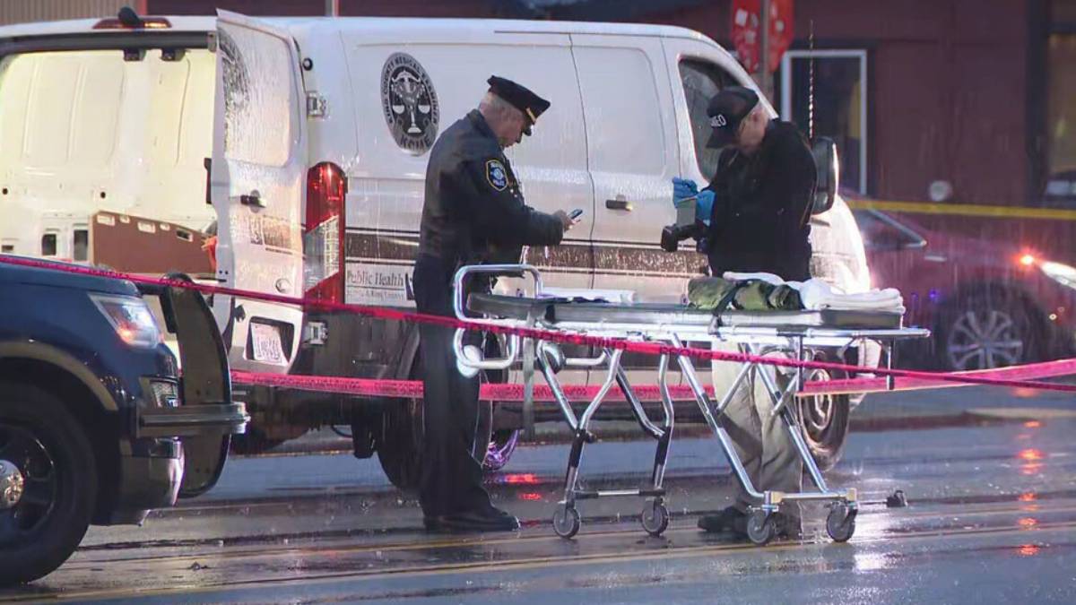 Emergency responders stand beside a wheeled stretcher at a taped-off crime scene, with a white van in the background and a police vehicle nearby.
