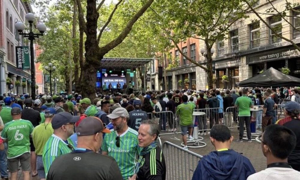 Crowd of spectators gathers along a city street to watch a stage event with performers in the distance under trees and buildings lining the avenue.