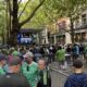 Crowd of spectators gathers along a city street to watch a stage event with performers in the distance under trees and buildings lining the avenue.