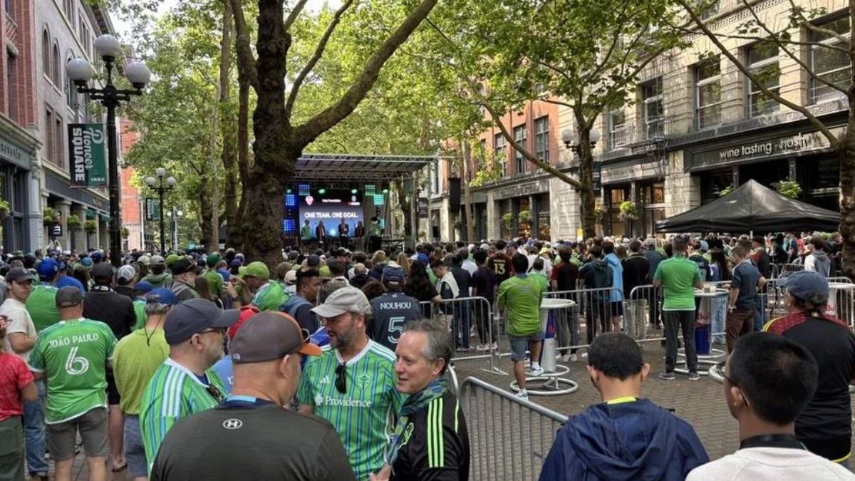 Crowd of spectators gathers along a city street to watch a stage event with performers in the distance under trees and buildings lining the avenue.
