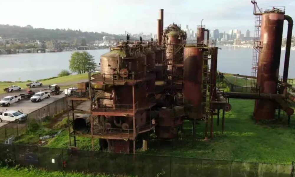Rusty, abandoned industrial complex with tall smokestacks and interconnected tanks by a river, city skyline in the distance.