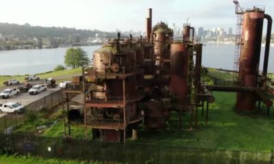 Rusty, abandoned industrial complex with tall smokestacks and interconnected tanks by a river, city skyline in the distance.