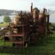 Rusty, abandoned industrial complex with tall smokestacks and interconnected tanks by a river, city skyline in the distance.